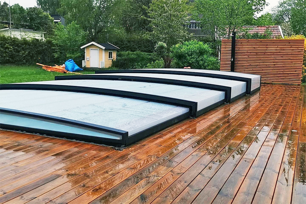 Aluna Poolüberdachung R1 in Flach-Ausführung auf nasser Holzterrasse, mit Garten im Hintergrund