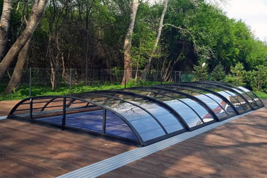 Flache Poolüberdachung Aluna Atlanta mit transparenten Segmenten auf Holzterrasse, umgeben von Bäumen