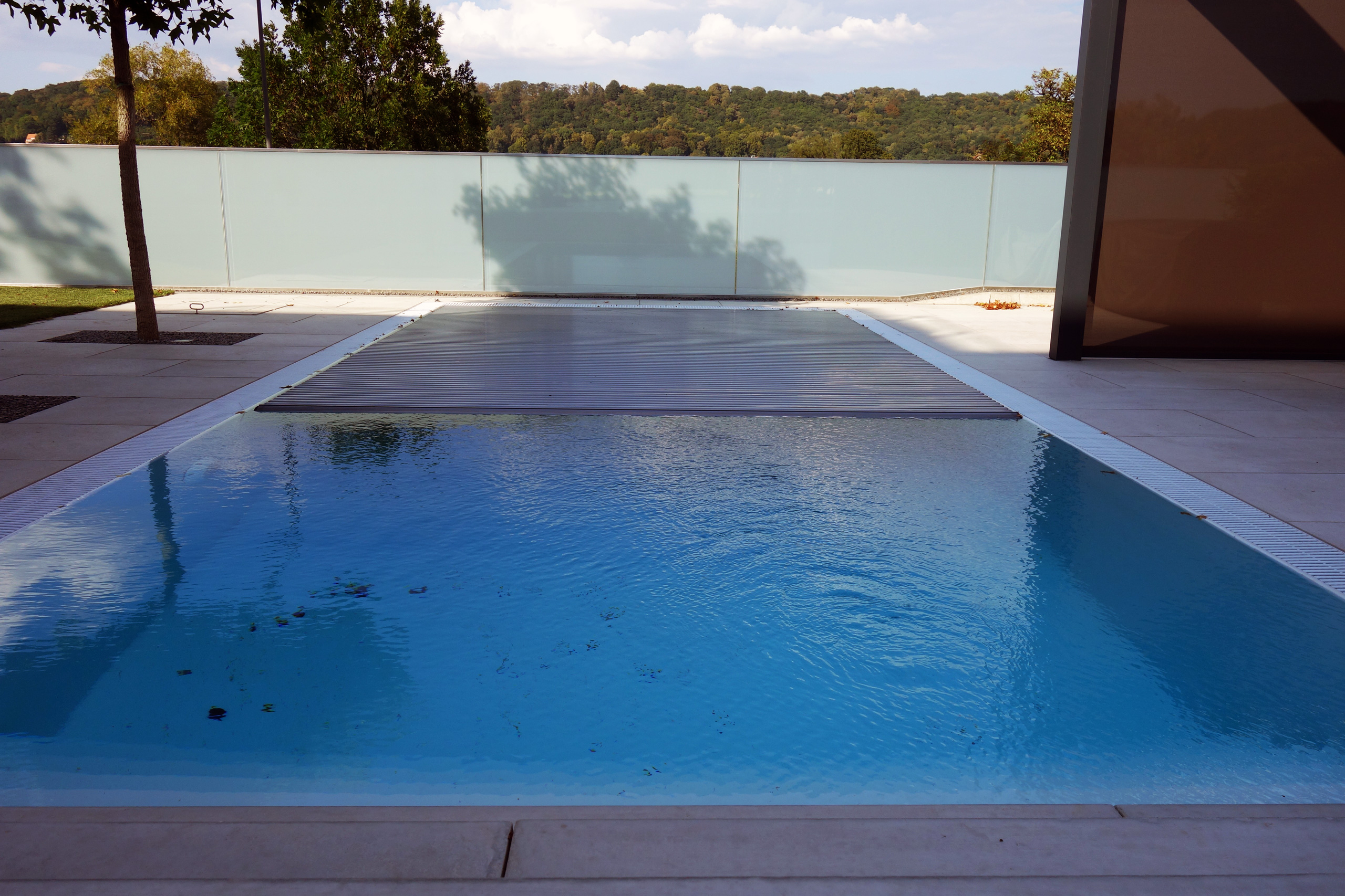 Überlaufpool mit blauer Folie, teilweise abgedeckt von einem Unterflurrolladen, umgeben von hellen Fliesen.