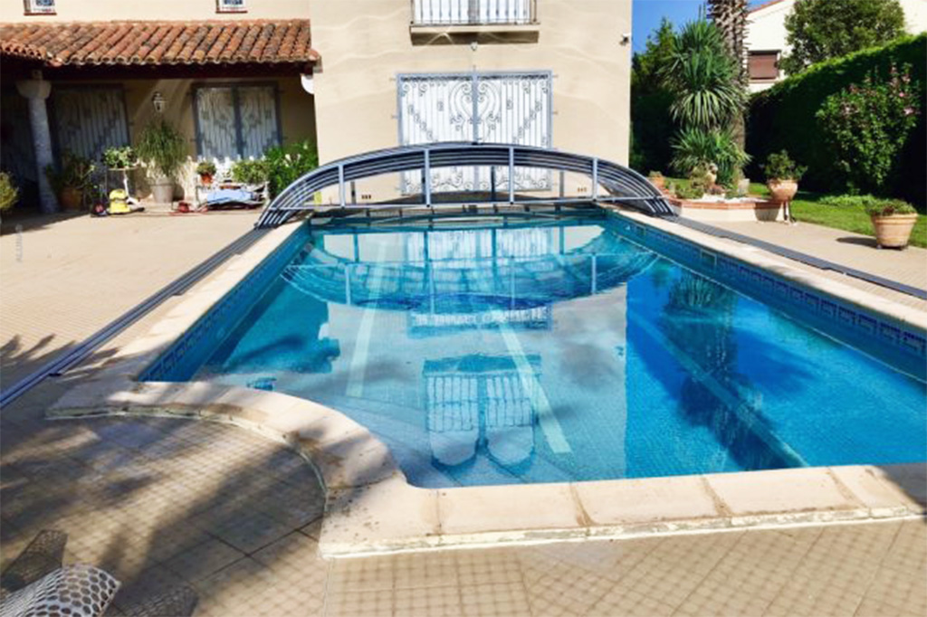 Flache Poolüberdachung Aluna Atlanta auf Schienen, teilweise über den Pool geschoben, mit Reflexionen im Wasser