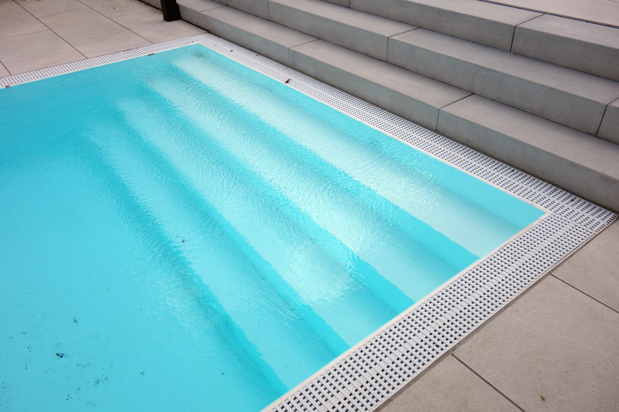 Gerade Treppe und Überlaufrinne eines Überlaufpools mit hellblauer Folie und grauen Fliesen.