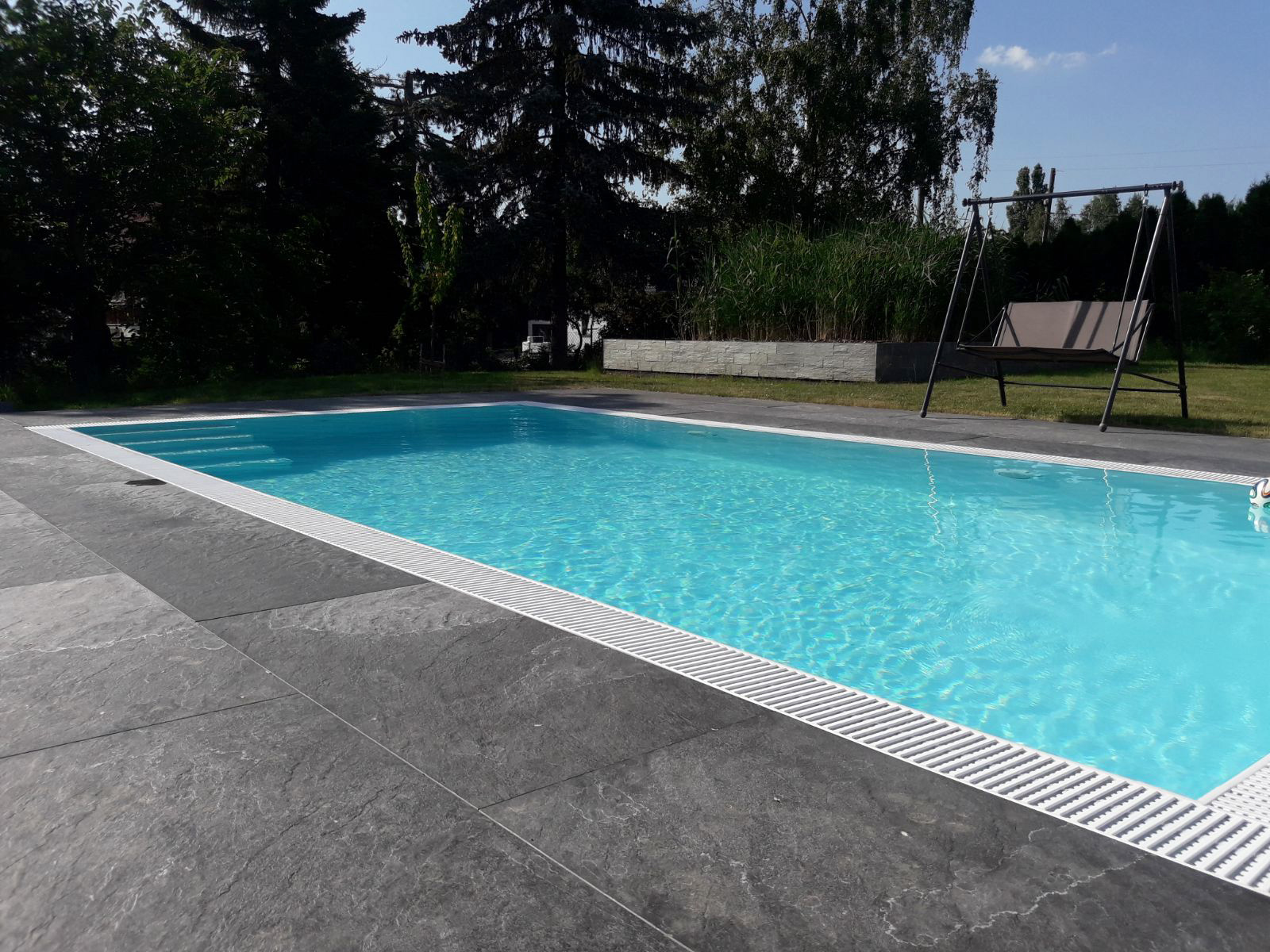 Weißer Albixon G2 Polypropylen Pool mit Ecktreppe und Überlaufrinne, umgeben von dunklen Steinplatten.