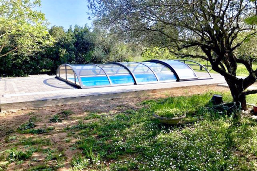 Flache Poolüberdachung Aluna Atlanta mit transparenten Segmenten und dunklem Rahmen in Gartenumgebung