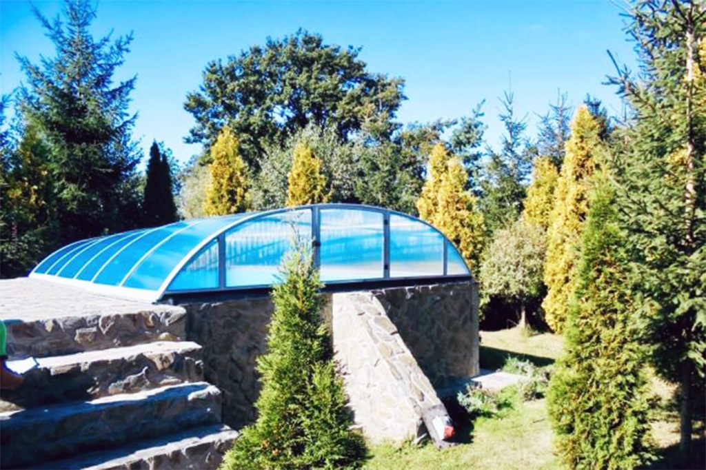 Flache Poolüberdachung Aluna Atlanta mit blauer Transparenz auf erhöhtem Pool in grüner Gartenlandschaft