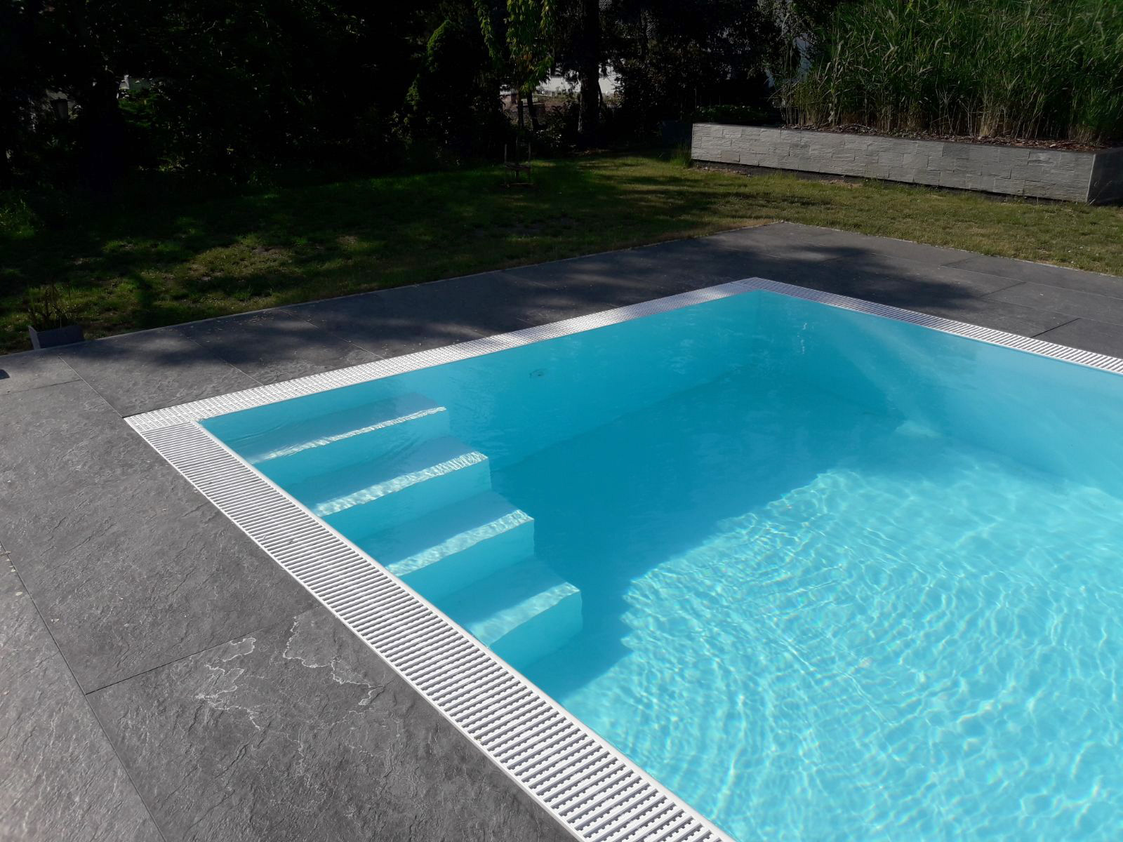 Weißer Albixon G2 Polypropylen Pool mit Ecktreppe und Überlaufrinne, umgeben von dunklen Steinplatten.